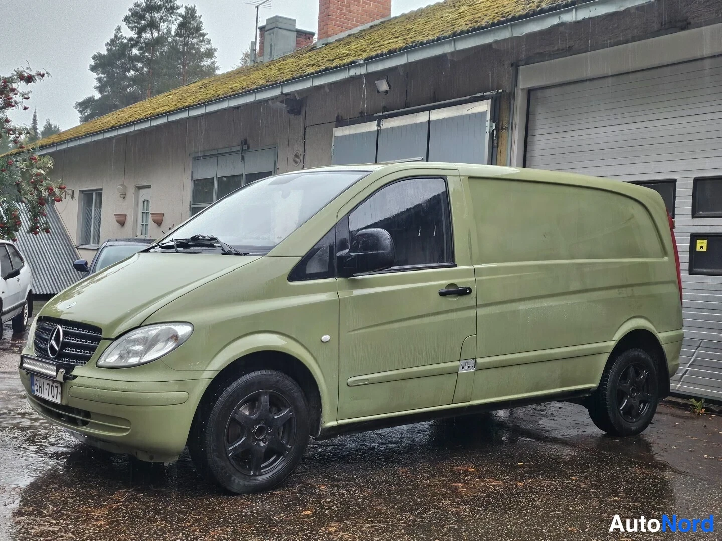 Etuviisto, nokka vasemmalle: Vihertävän ruskeanvärinen Mercedes-Benz Vito -pakettiauto etuviistosta vasemmalta; rekisterikilpi, mustat vanteet, lisävalo puskurissa, märkä piha ja v