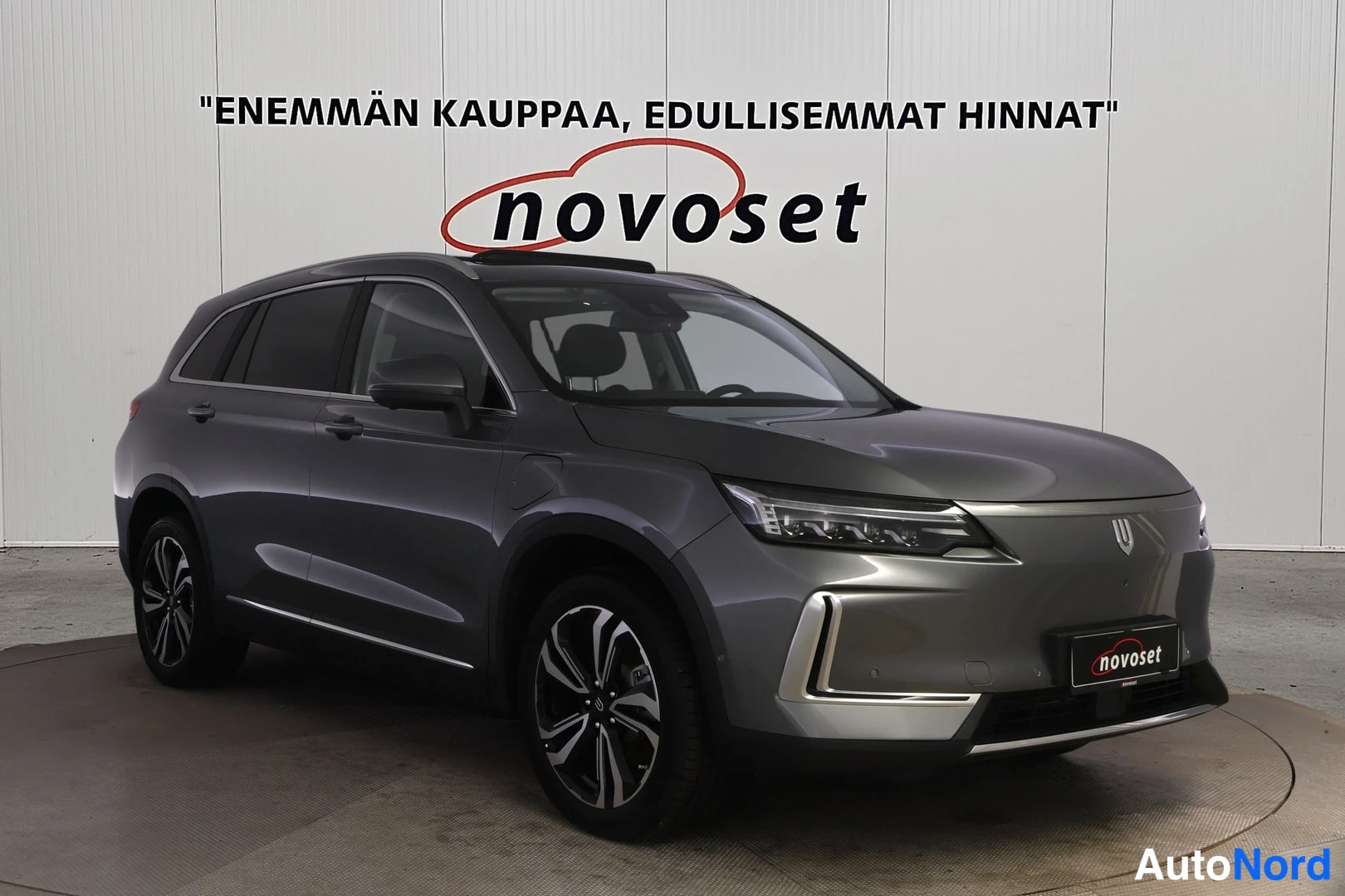 Etuviisto, nokka oikealle: Harmaa KYWELL-nimellä mainostettu farmarityyppinen sähköauto kolmesta neljänneksestä edestä oikealta kuvattuna autonmyyntihallissa.
