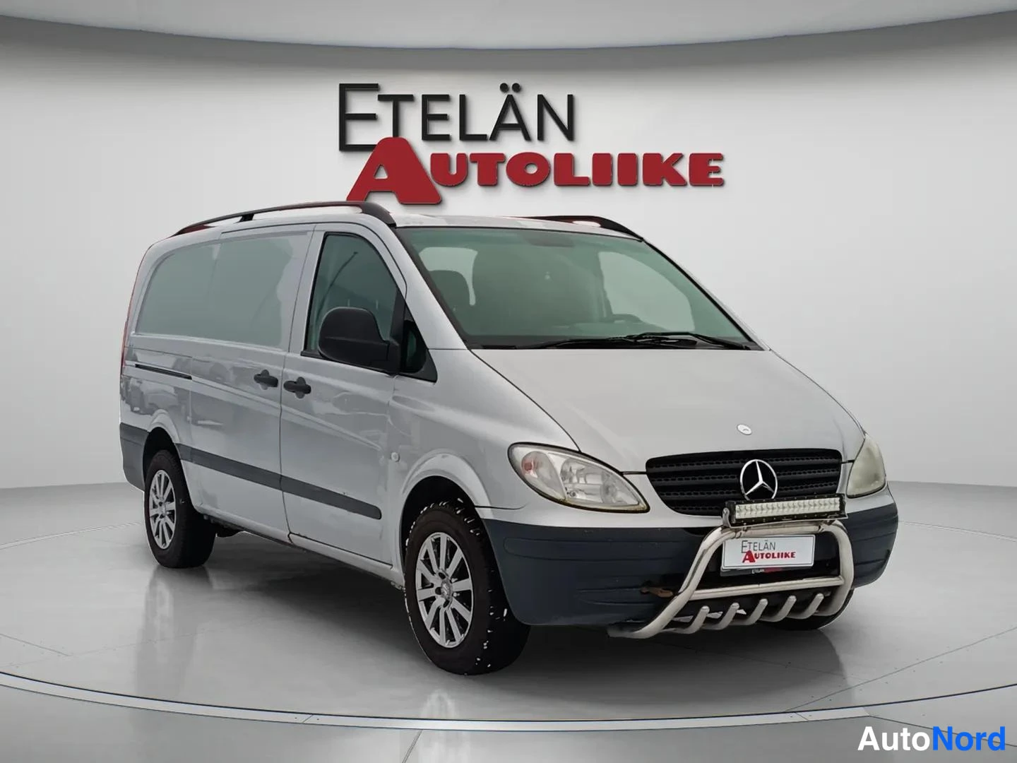 Etuviisto, nokka oikealle: Mercedes-Benz Vito -pakettiauto edestä oikealta kolmasosan kulmakuvassa, hopeisenä, kattotelineet ja etupuskurin suojarUNKO. Grille near right edge with