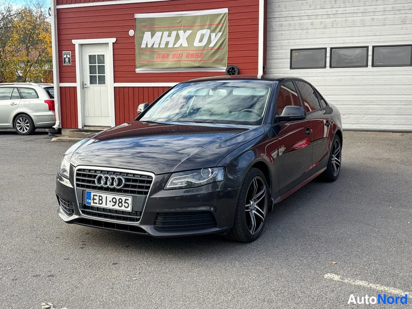Etuviisto, nokka vasemmalle: Tummanharmaa Audi A4 -sedan etuviistossa, rekisterikilpi, audi-merkki etusäleikössä ja tummat vanteet.