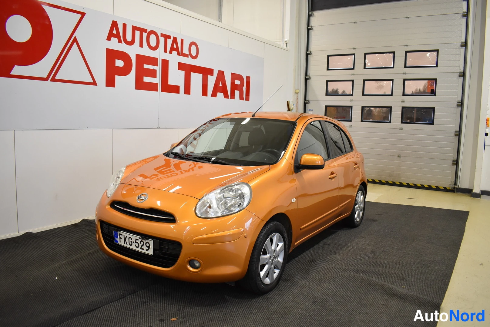 Etuviisto, nokka vasemmalle: Oranssi Nissan Micra -henkilöauto nähty edestä vinosti autotallin sisällä, rekisterikilpi.