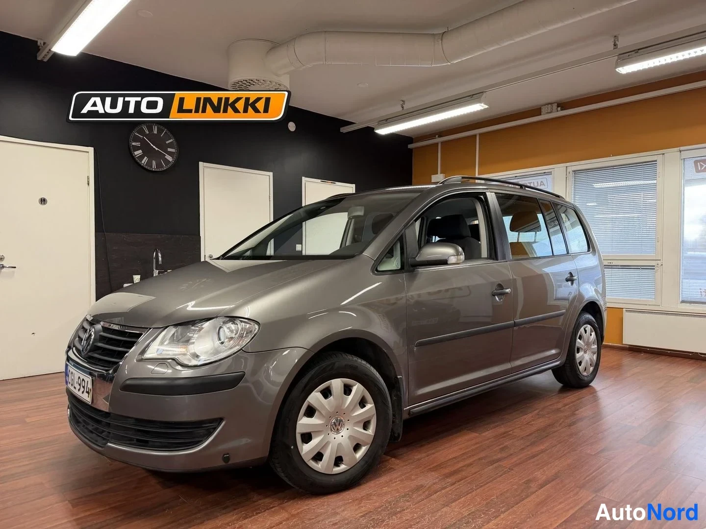 Etuviisto, nokka vasemmalle: Hopeanharmaa Volkswagen Touran -henkilöauto auton sisätiloissa, etukulmasta vasemmalta kuvattuna. Etusäleikön VW-merkki, sumukatto ja lisävalot näkyvät