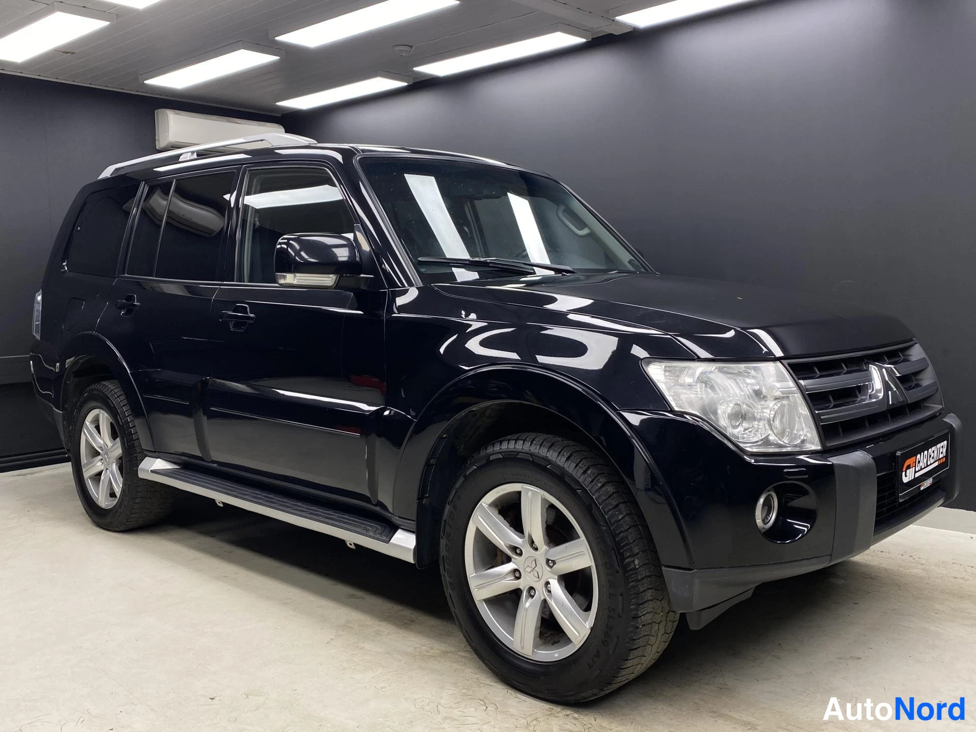 Etuviisto, nokka oikealle: Musta Mitsubishi Pajero etukulmasta oikealta; näkyvillä ajovalo, etusäleikkö, ovien kahvat, kattoteline ja sivustepit.