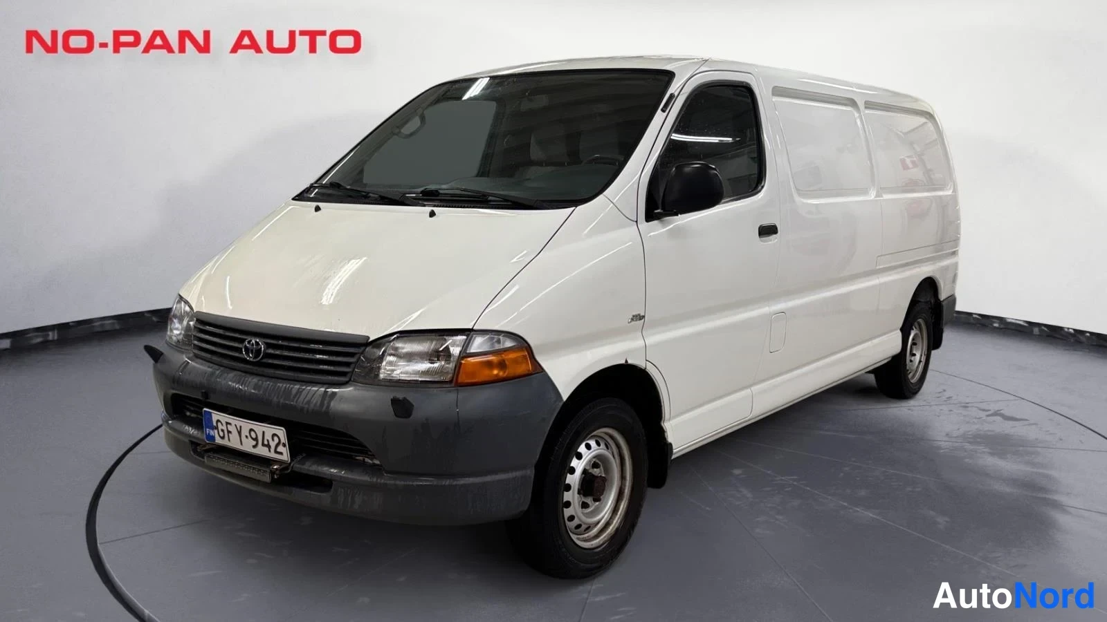 Etuviisto, nokka vasemmalle: Toyota Hiace -pakettiauto etuviistosta valkopanoraamassa, rekisterikilpi.