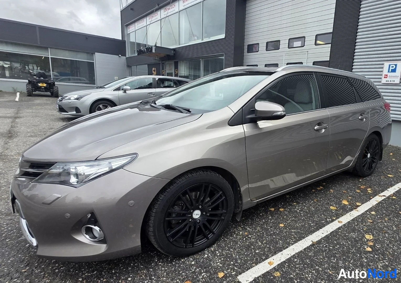 Etuviisto, nokka vasemmalle: Tummanharmaan Toyota Auris Touring Sports -farmarin etkolänurkkakuva; mustat kevytmetallivanteet, kattokaiteet ja klaarit ajovalot näkyvät pysäköintial