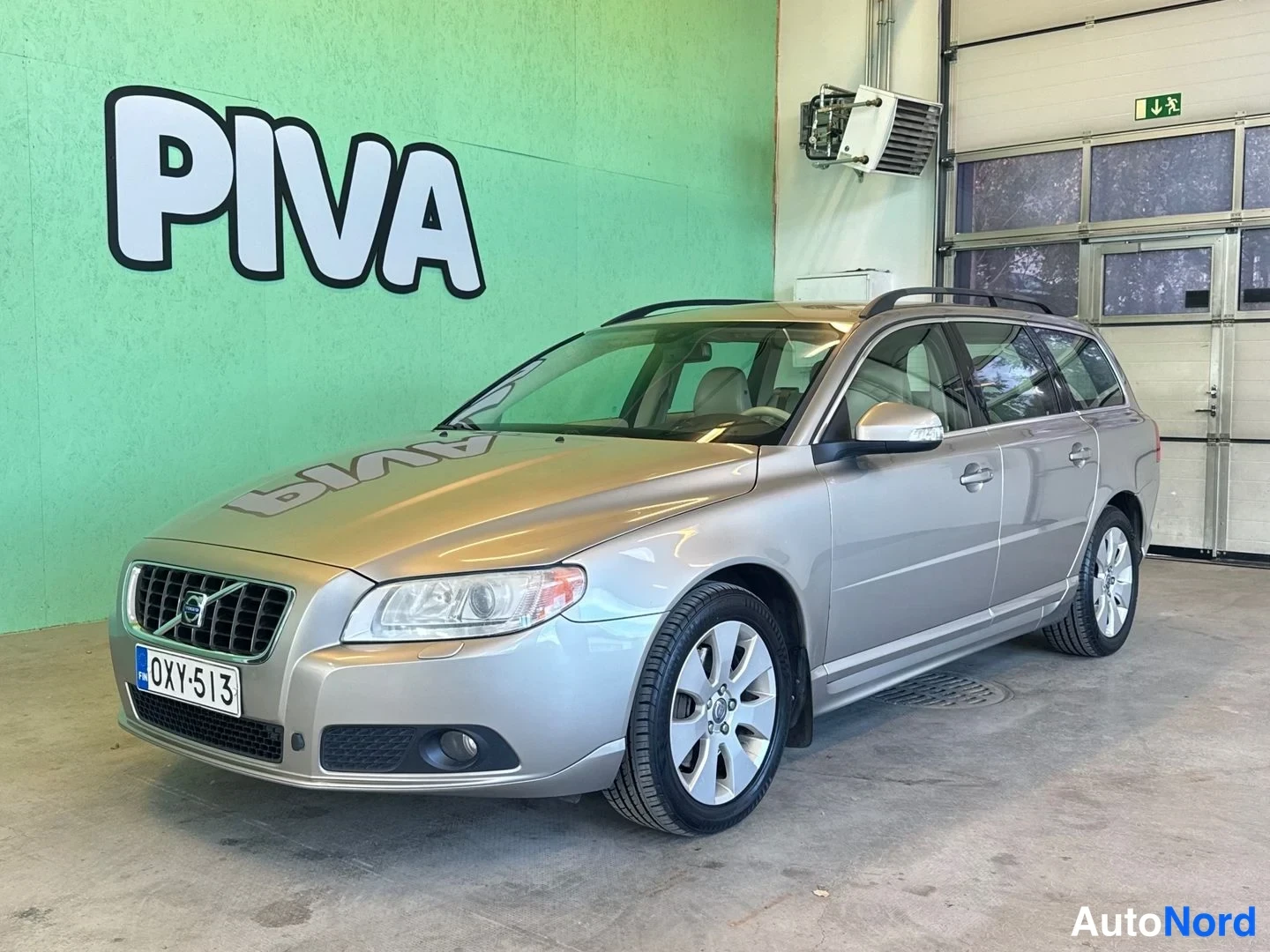 Etuviisto, nokka vasemmalle: Volvo V70 -farmari kylki ja keula kuvakulmassa keula vasemmalla, hopeanvärinen kori, kattokaiteet, rekisteritunnus, PIVA-mainos seinällä.